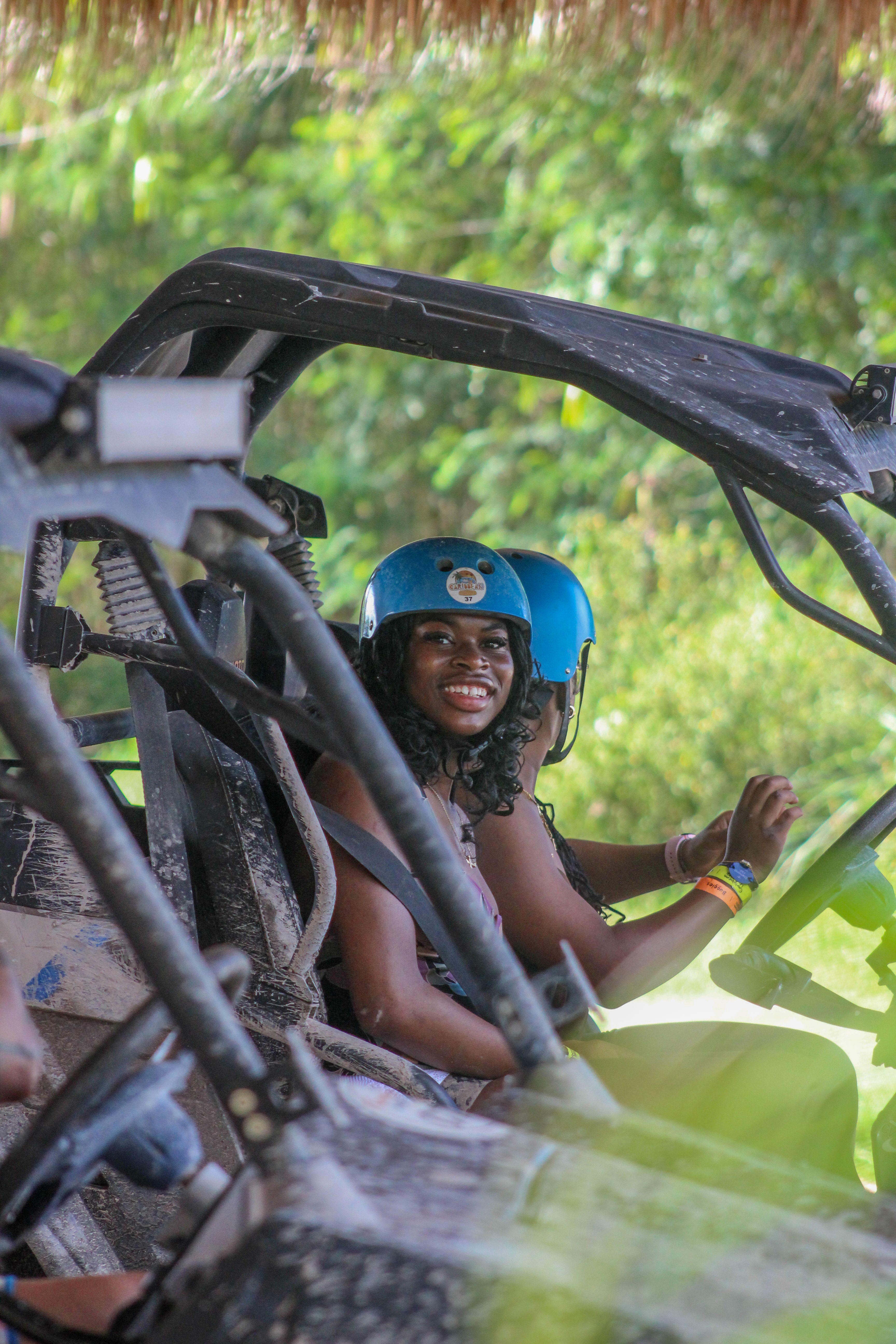 Smiling person in a blue helmet seated in a muddy off-road buggy/UTV against lush tropical greenery, ready for an outdoor adventure