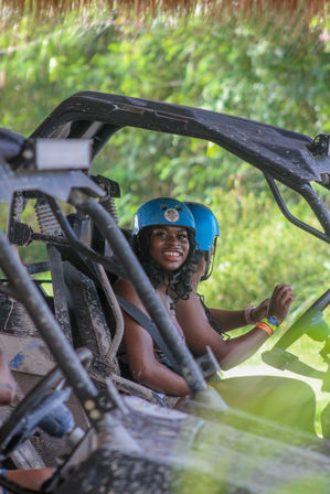 Smiling person in a blue helmet seated in a muddy off-road buggy/UTV against lush tropical greenery, ready for an outdoor adventure