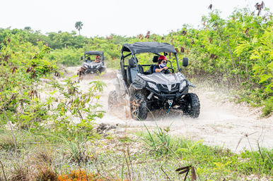 Two off-road side-by-side UTVs splashing through a muddy tropical trail; lead driver in a red helmet and bandana steers as water sprays, surrounded by dense green scrub and palms.