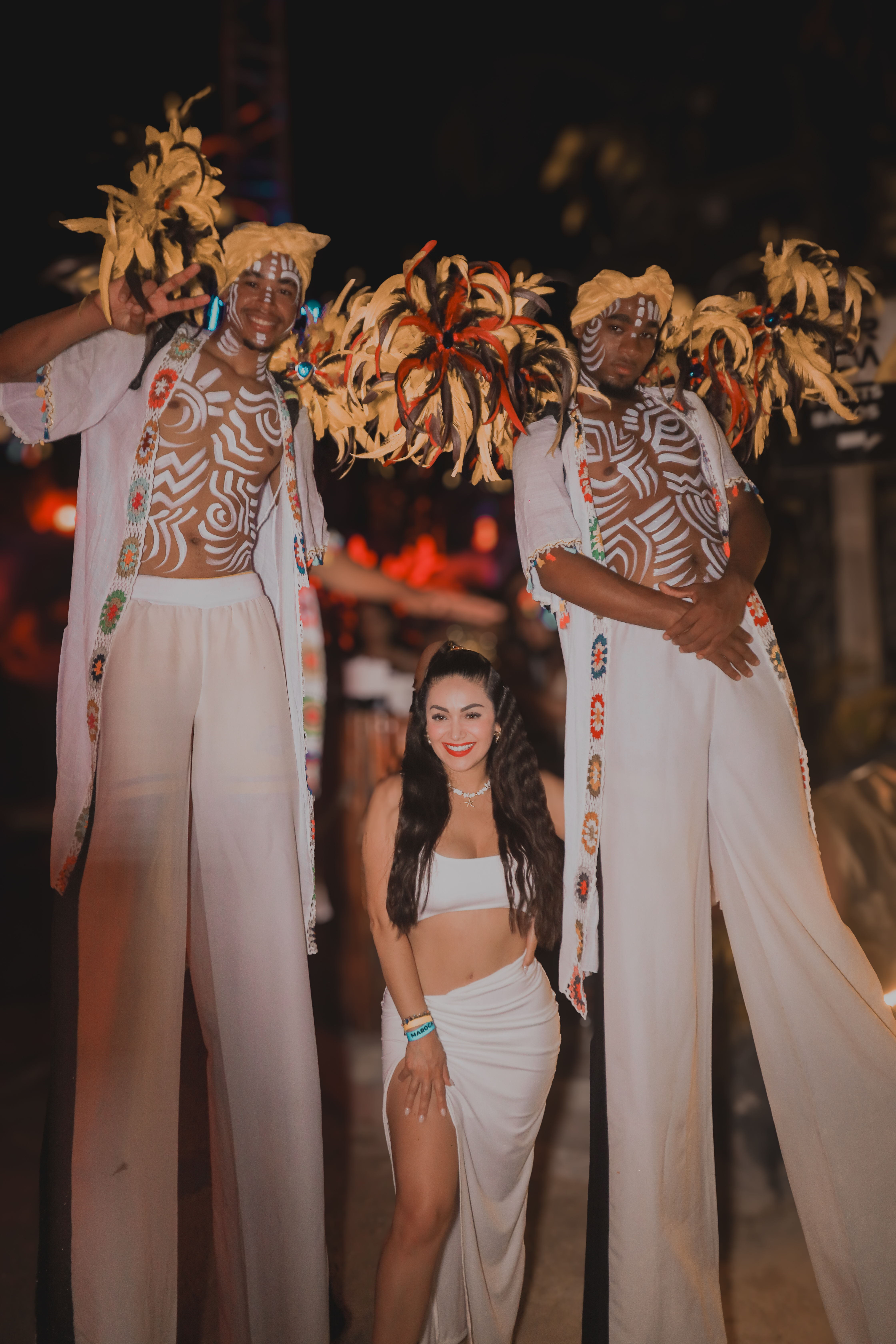 Nighttime tropical festival: two tall stilt performers with colorful feather headdresses and white body paint flank a smiling woman in a white crop top and flowing skirt.