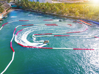 Aerial view of jet skis racing a buoy-lined watersports course in a turquoise tropical lagoon with palm trees and sunlit splashing trails.