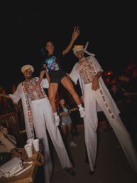 Smiling woman lifted between two stilt walkers with white body paint and embroidered robes at a lively nighttime outdoor party or festival, crowd and drinks visible in the background.