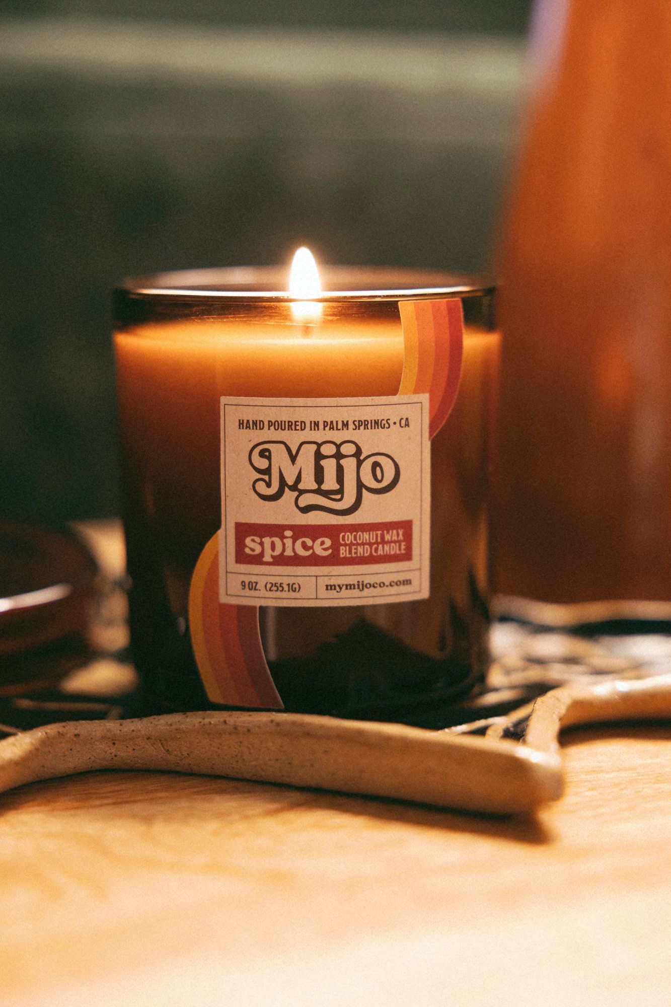 Cozy close-up of a lit amber-glass spice-scented candle with a warm flame and retro label resting on a wooden surface with ceramic decor