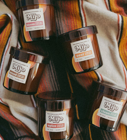 Five amber-glass scented candles labeled bloom, zest, wood, spice and clean arranged on a striped Southwestern blanket, Palm Springs desert-chic vibe.
