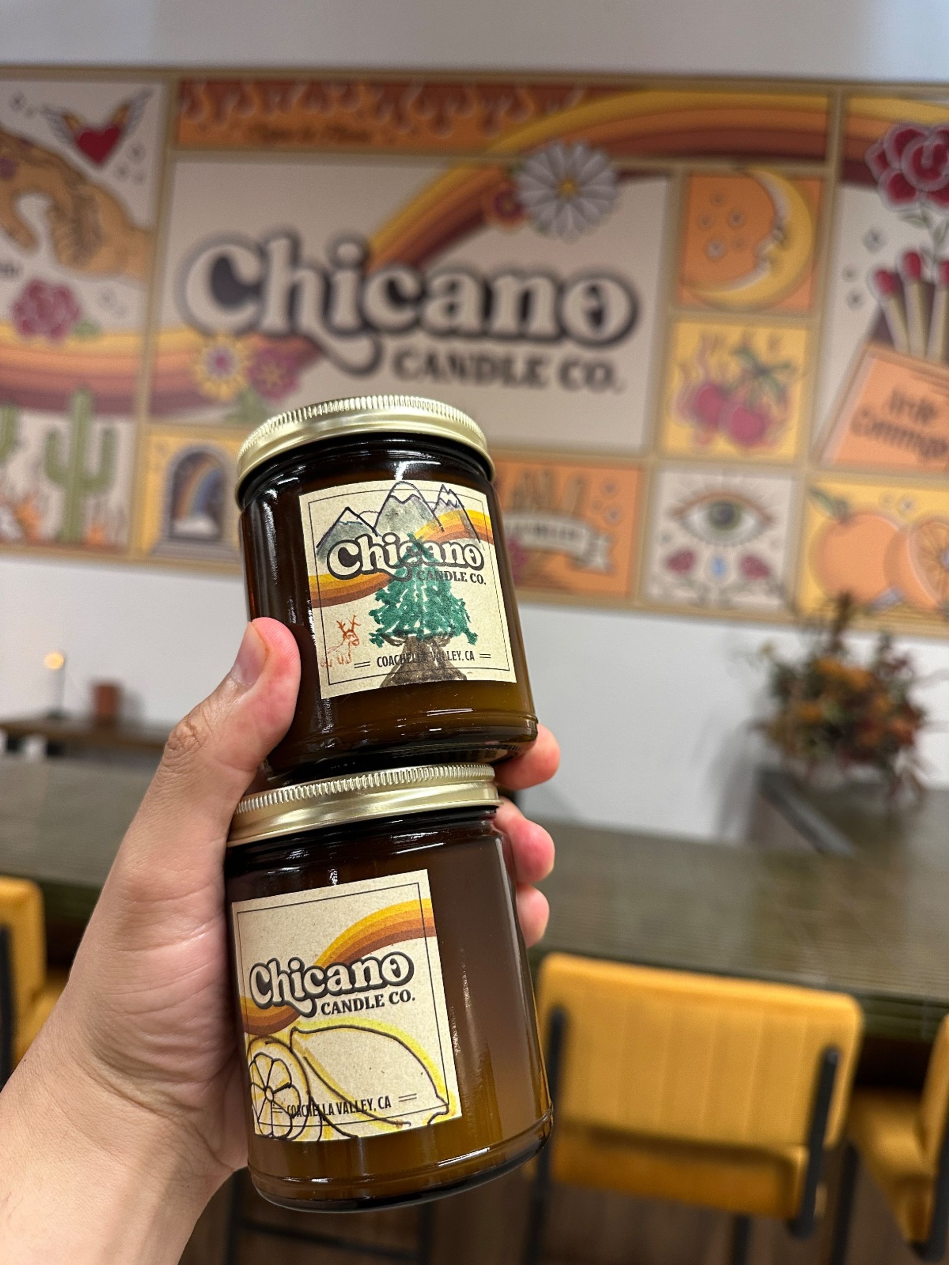 Hand holding two amber glass candle jars with retro sunset and tree labels, blurred colorful tiled mural and wooden chairs in a cozy candle shop interior.