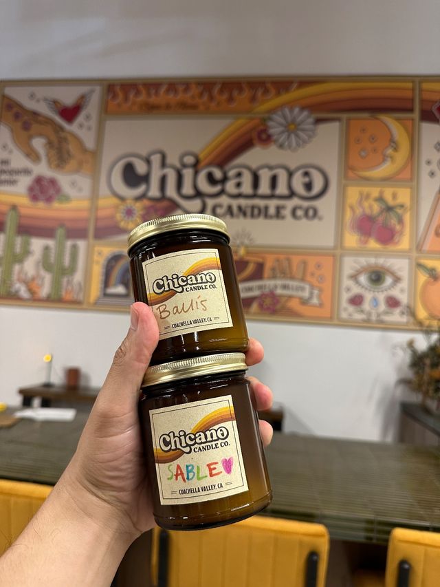Hand holding two amber glass jar candles with retro rainbow labels in a colorful desert‑themed shop interior, mural featuring cacti, sun, moon and floral motifs.