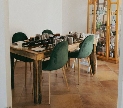 Cozy dining room scene with a rustic wooden table covered in metal cups, trays and small bottles, surrounded by green velvet chairs beside a glass-front liquor cabinet on warm terracotta tile flooring.
