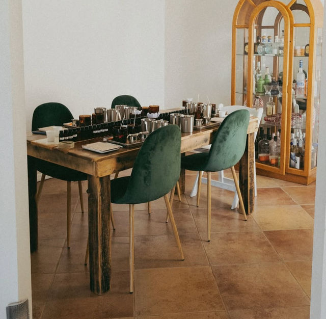 Cozy dining room scene with a rustic wooden table covered in metal cups, trays and small bottles, surrounded by green velvet chairs beside a glass-front liquor cabinet on warm terracotta tile flooring.