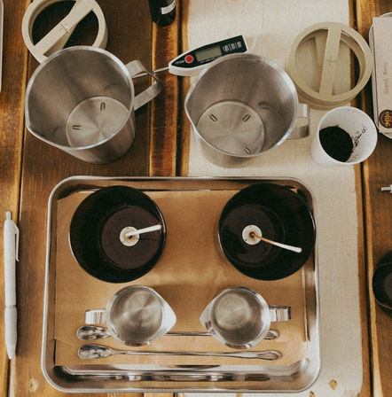 Overhead shot of a DIY candle-making workspace on a wooden table: metal tray with two black jars and wicks, stainless steel pour pitchers, spoons, digital thermometer and a small cup of dark granules.