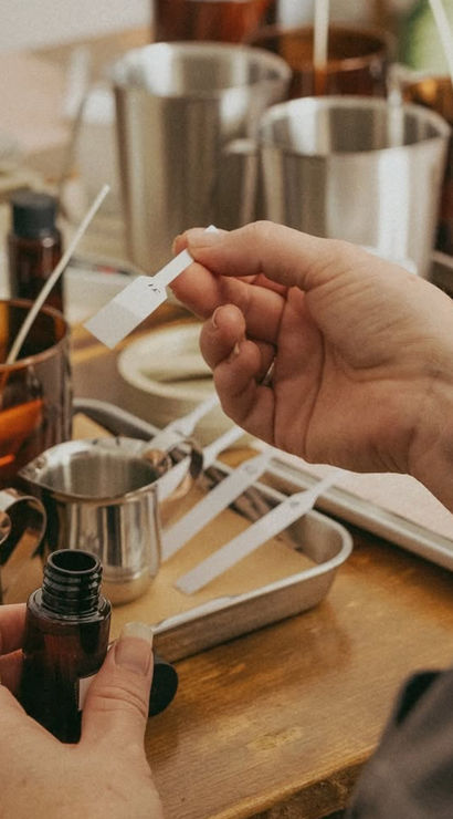 Hands holding a scent strip and amber dropper bottle at an artisanal fragrance-blending workshop with stainless jugs, trays and testing strips on a wooden table.