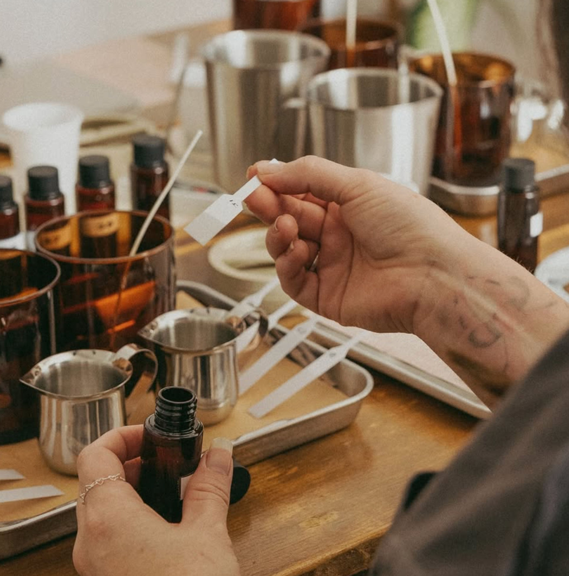 Hands holding a scent strip and amber dropper bottle at an artisanal fragrance-blending workshop with stainless jugs, trays and testing strips on a wooden table.