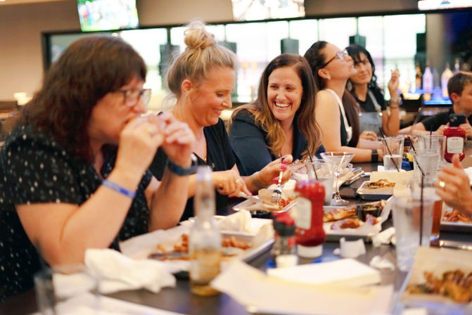 Smiling group of friends sharing plates and drinks at a busy bar counter in a casual restaurant, laughing over appetizers and cocktails.