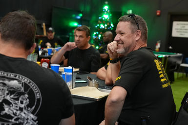 Smiling man in a black shirt chats with colleagues at an indoor holiday gathering, trays and blue cups on the table and a lit Christmas tree in the background.