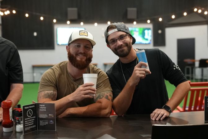 Two smiling men at an indoor sports bar counter holding drinks—one with a white cup, the other with a blue can—string lights overhead and a green turf play area with TV screens blurred in the background.
