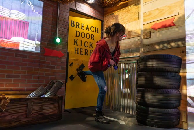 Participant in a red hoodie kicking a yellow "BREACH DOOR KICK HERE" panel in an indoor interactive obstacle course, surrounded by stacked tires, corrugated metal walls, red arrows and a green signal light.