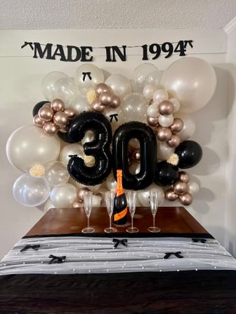 Indoor 30th birthday backdrop with large black '30' balloons and 'MADE IN 1994' banner, rose-gold, black, white and clear balloon garland, champagne bottle and four flutes on a black-and-white tulle-decorated table.