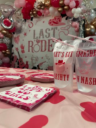 Pink Western-themed bachelorette party setup with 'Last Rodeo' backdrop, pink and gold balloon arch, heart-print tablecloth, themed napkins, plates and drink pouches with straws.