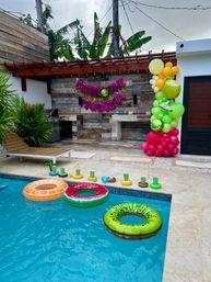 Bright tropical backyard pool party setup with three fruit-themed inflatable rings (kiwi, watermelon, orange) floating in a turquoise pool, poolside lounge and tiled outdoor bar under a wooden pergola, pink balloon-letter banner reading “GIMME ONE MARGARITA” and a lime-and-yellow balloon column with tropical plants.