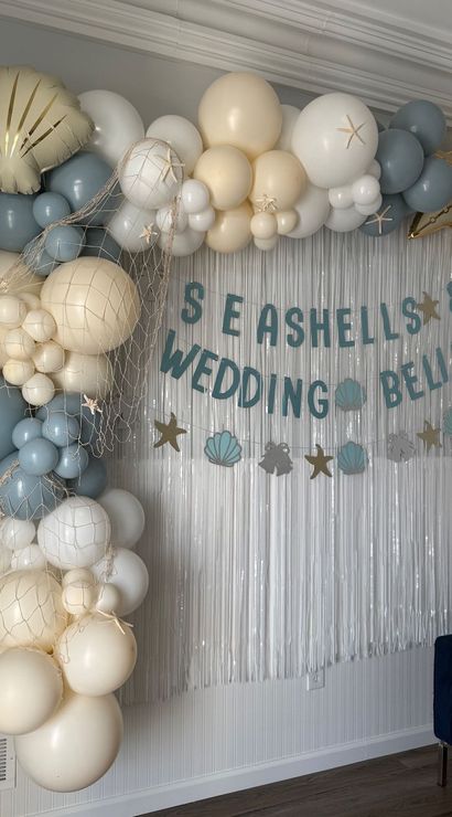 Coastal indoor wedding backdrop with cream and blue balloon arch, gold star and shell foil balloons, fishing-net accents and a blue-letter "Seashells & Wedding Bells" banner over a silver fringe curtain.