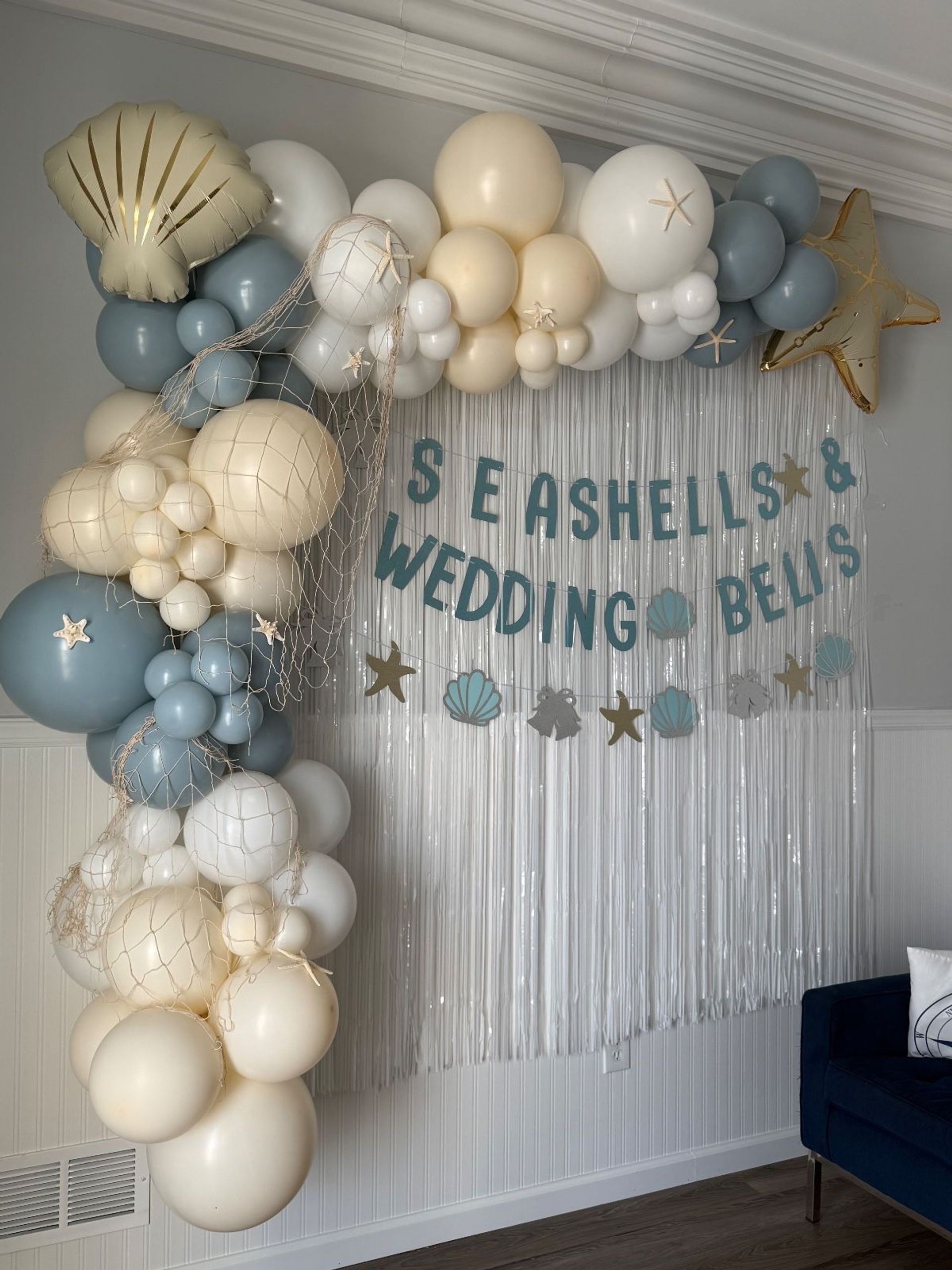 Coastal indoor wedding backdrop with cream and blue balloon arch, gold star and shell foil balloons, fishing-net accents and a blue-letter "Seashells & Wedding Bells" banner over a silver fringe curtain.