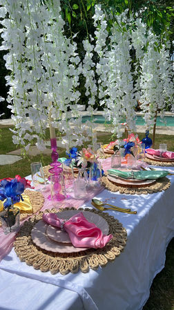 Poolside outdoor tablescape under cascading white wisteria with woven placemats, pastel plates and napkins, colorful glassware and gold flatware in a bright summer garden setting