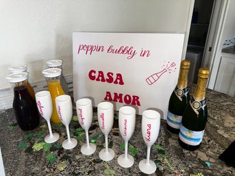 Home mimosa bar on a granite kitchen counter with two bottles of sparkling wine, carafes of orange and cranberry juice, six white personalized champagne flutes, confetti, and a large sign reading poppin bubbly in CASA AMOR.
