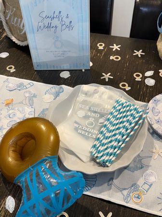Beach-themed bachelorette party table with “Seashells & Wedding Bells” sign, blue-striped paper straws, white shell plate, inflatable drink holder and scattered seashell confetti on a dark wood table.