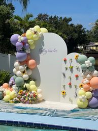 Outdoor poolside baby shower backdrop with white arched panels, pastel balloon garlands, sunflower accents and colorful floral arrangements under blue sky and palm trees