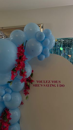 Light-blue balloon arch with bright pink floral garland framing a cream sign that reads "VOULEZ VOUS SHE'S SAYING I DO" in front of a teal metallic fringe backdrop — indoor bridal shower display.