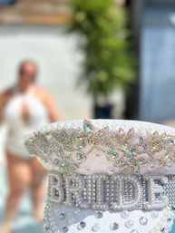 Sparkling white bridal cap covered in pearls and rainbow rhinestones with 'BRIDE' lettering, photographed poolside with a blurred person and tropical greenery in the background.
