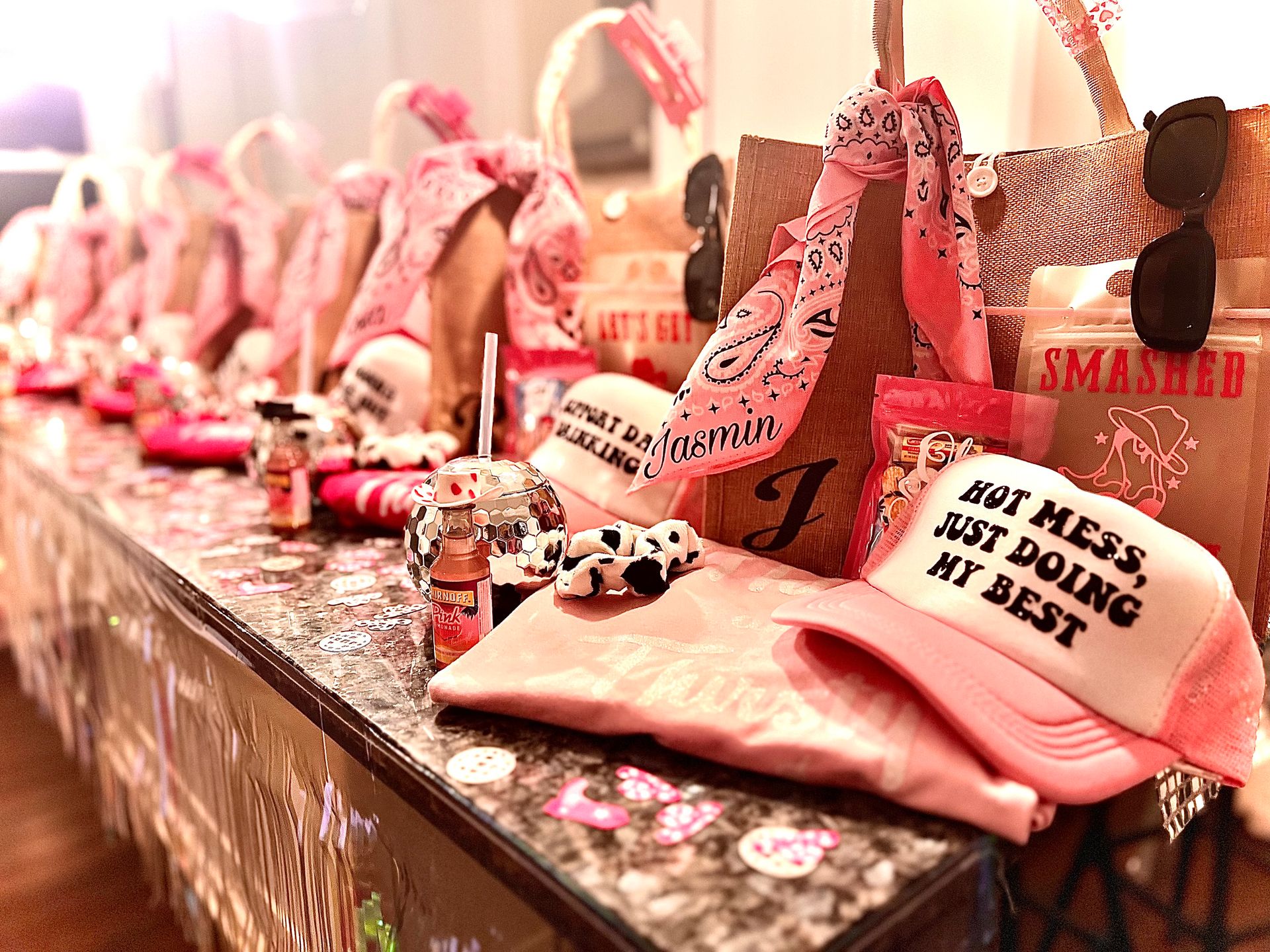 Pink bachelorette-style party favor display on a marble countertop: a row of gift bags with personalized bandanas, sunglasses, snack packs, disco-ball cups and a pink trucker hat reading 'HOT MESS, JUST DOING MY BEST'.