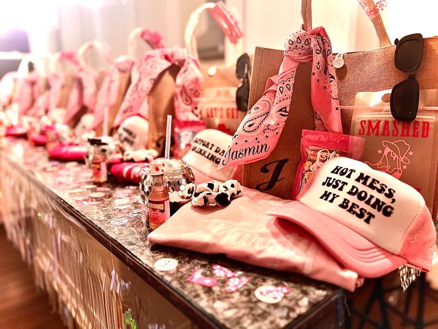 Pink bachelorette-style party favor display on a marble countertop: a row of gift bags with personalized bandanas, sunglasses, snack packs, disco-ball cups and a pink trucker hat reading 'HOT MESS, JUST DOING MY BEST'.