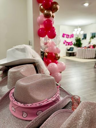 Sparkly pink cowboy hats with paisley bandanas in the foreground, tall pink and rose-gold balloon garland and festive indoor party setup with table and chandelier in the background.