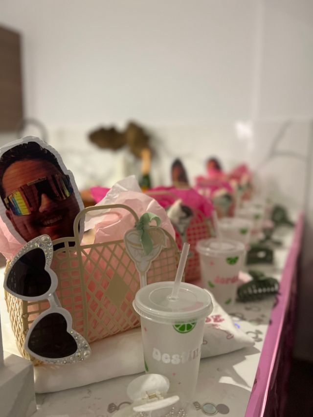 Row of pink party favor baskets on a table with tissue paper, heart-shaped sunglasses, novelty face cutouts, and plastic cups with lids and straws — playful party favor table setup.