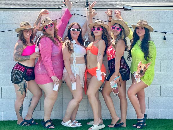 Six friends in colorful swimsuits, mesh cover-ups and straw hats posing with sunglasses and drinks against a cinderblock wall at a sunny backyard pool party.