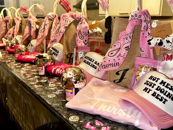 Pink-themed party favor display on a kitchen counter: lined gift bags with pink bandanas, novelty caps with playful slogans, mini disco balls, drink mixers and confetti for a bachelorette or girls' night celebration.