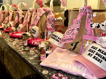 Pink bachelorette party favors lined up on a kitchen island: burlap tote bags tied with pink bandanas, mirrored disco-ball cups with straws, slogan trucker hats, folded pink tees, confetti and mini drink bottles.