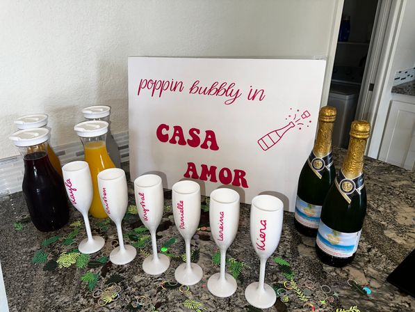 At-home mimosa bar on a granite kitchen counter with a decorative sign reading 'poppin bubbly in CASA AMOR', two bottles of sparkling wine, juice carafes, six personalized white champagne flutes and tropical confetti