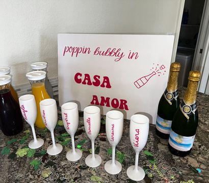 At-home mimosa bar on a granite kitchen counter with a decorative sign reading 'poppin bubbly in CASA AMOR', two bottles of sparkling wine, juice carafes, six personalized white champagne flutes and tropical confetti