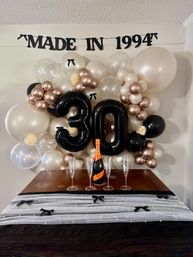 Elegant indoor 30th birthday backdrop with 'MADE IN 1994' banner, large black '30' balloons set in a pearl white, rose-gold and black balloon garland, and a table with a sparkling wine bottle and four champagne flutes.