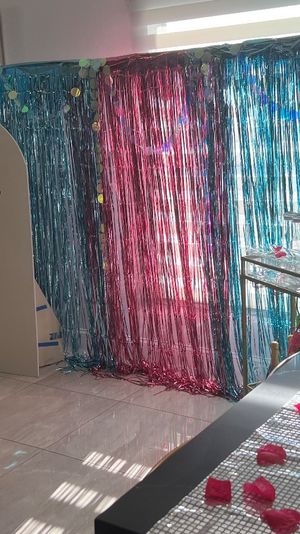 Modern home party setup with blue balloon column and disco balls beside a pink-and-blue metallic fringe photo backdrop, kitchen island lined with martini glasses and scattered pink rose petals.