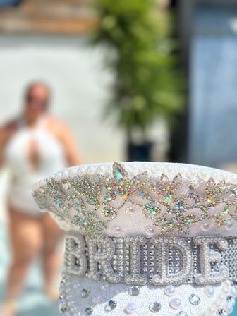 Close-up of a sparkling white bridal hat decorated with pearls and iridescent rhinestones spelling BRIDE, with a blurred poolside scene and person in a white swimsuit in the background.