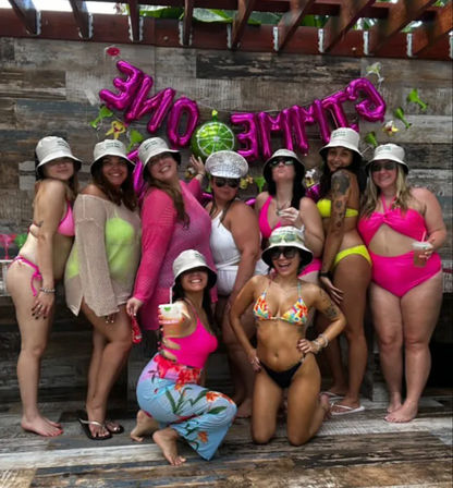 Group of friends in bright swimsuits and matching bucket hats posing on a tropical wood deck with pink celebration balloons and cocktails — lively poolside party vibe