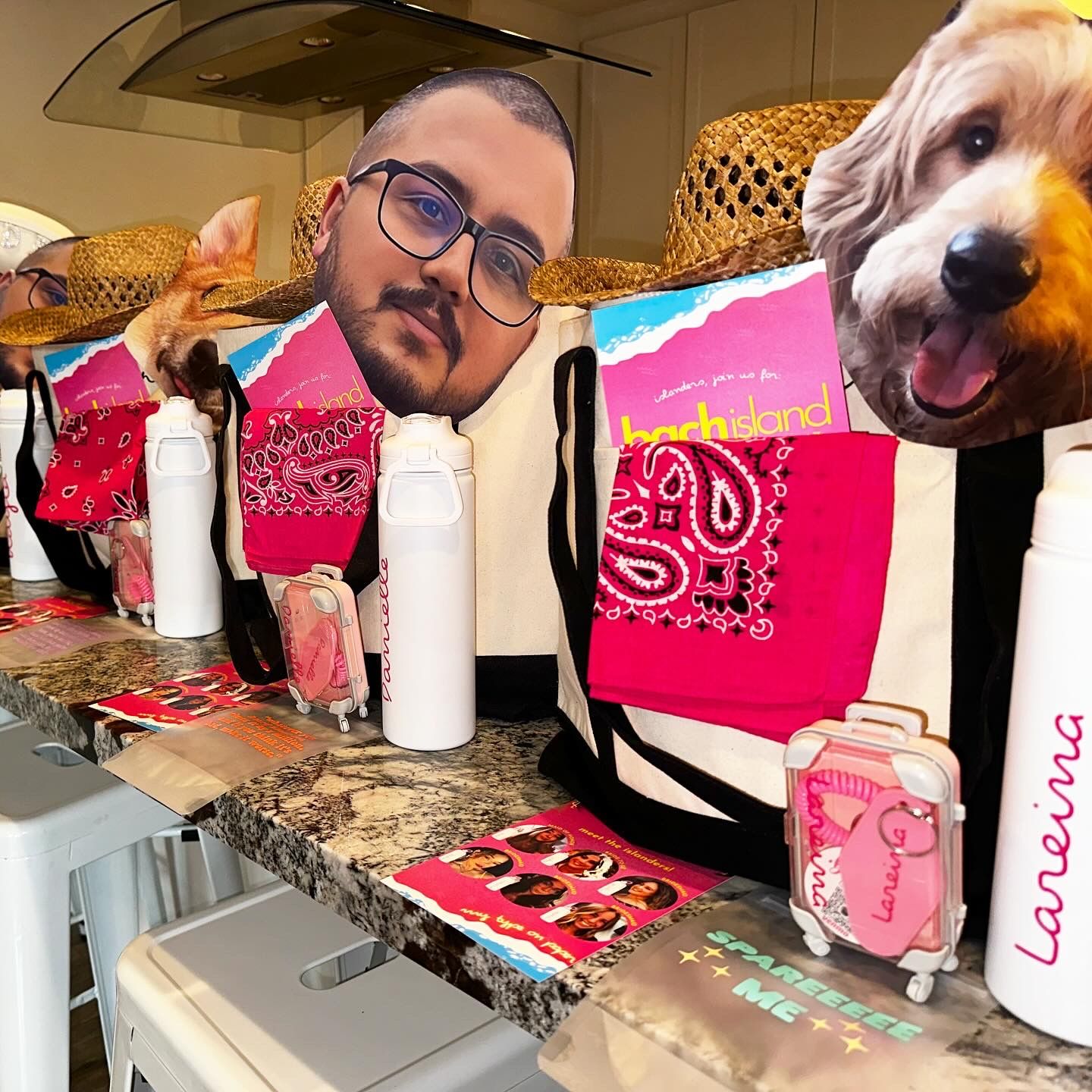 Bright beach-themed party favor lineup on a kitchen island: canvas tote bags with straw hats, oversized face cutouts (people and a dog), pink bandanas, white water bottles, mini pink luggage toys and colorful beach getaway brochures.