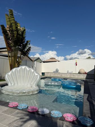 Sunny suburban backyard pool with a large inflatable white shell lounger, blue clear tubes floating, colorful shell toys on the stone deck, tall palm and white fence under a bright blue sky with puffy clouds