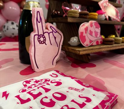 Pink-themed party dessert table with “LET’S GO GIRLS” napkins, champagne bottle, playful middle-finger hand cutout, cupcakes and macarons on a wooden tiered stand and a pink balloon garland backdrop.