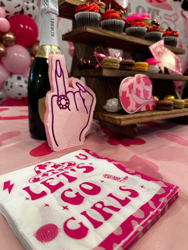 Pink-themed party dessert table with “LET’S GO GIRLS” napkins, champagne bottle, playful middle-finger hand cutout, cupcakes and macarons on a wooden tiered stand and a pink balloon garland backdrop.