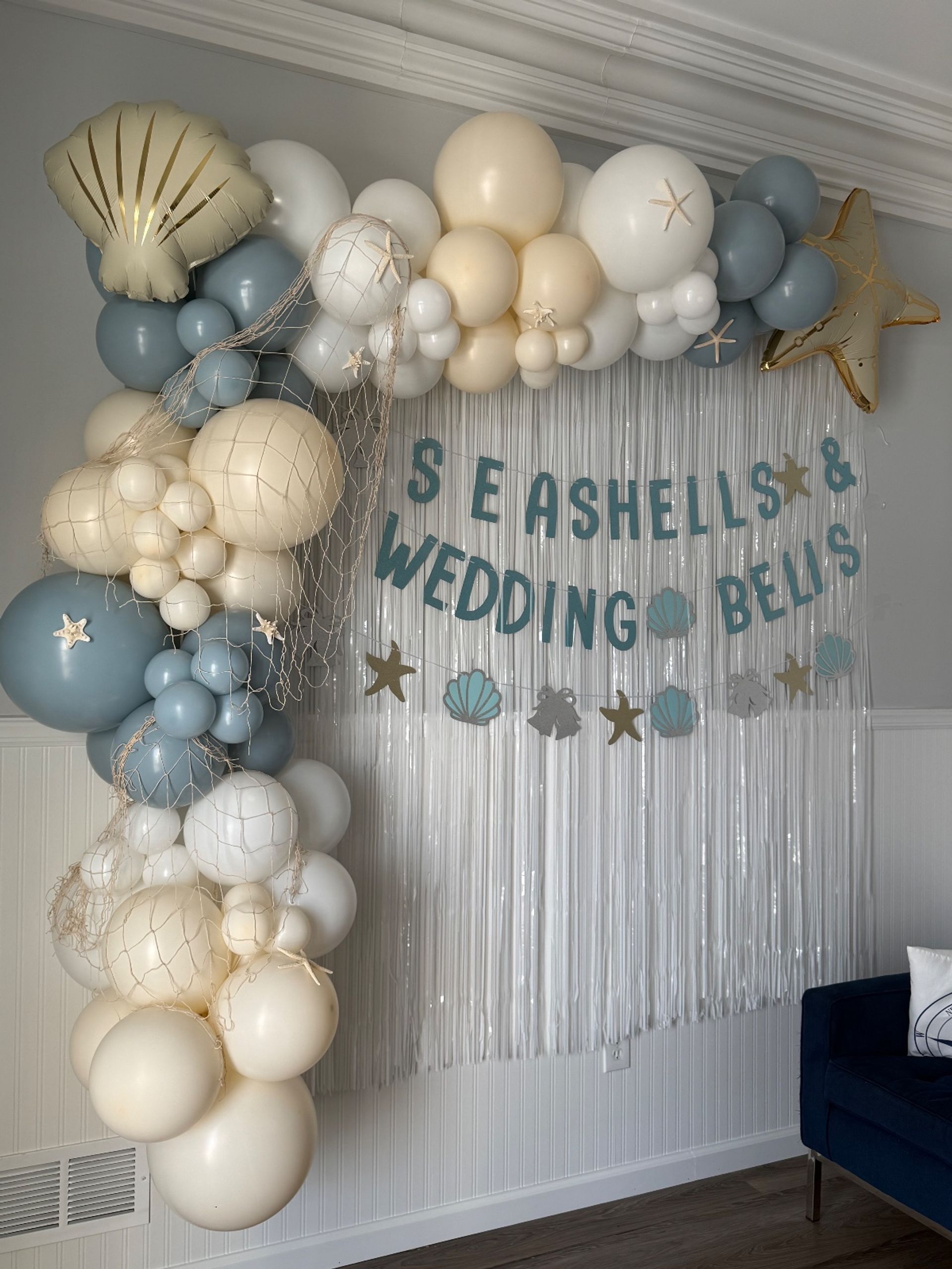 Indoor beach-themed wedding backdrop with cream, white and dusty-blue balloon arch, gold shell and star balloons, fishing-net accents and metallic fringe curtain with a 'Seashells & Wedding Bells' banner.