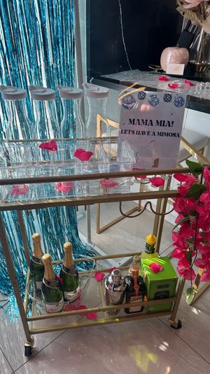 Gold bar cart mimosa station with champagne bottles, mixers, clear carafes and scattered pink flower petals, blue metallic fringe backdrop and a sign reading "MAMA MIA! LETS HAVE A MIMOSA" for a festive brunch setup