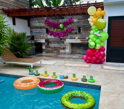 Backyard pool party setup with blue pool, colorful fruit-shaped inflatable floats (orange, watermelon, kiwi), margarita-themed banner, vibrant balloon column, wooden pergola and tropical plants on an outdoor patio.